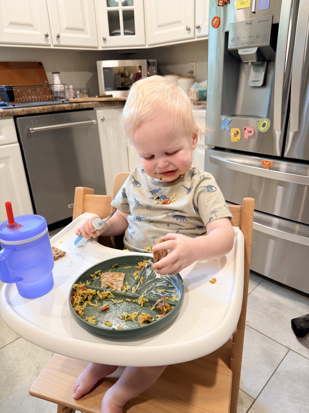 A smiling toddler self-feeding rice and finger foods in a high chair